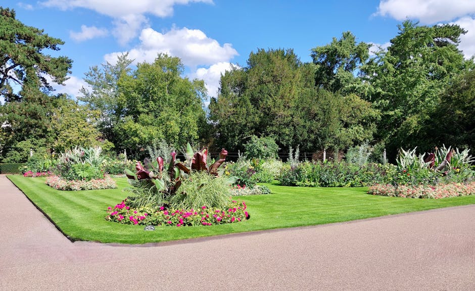 A well-maintained garden featuring lush green grass and neatly edged flower beds filled with vibrant pink, purple, and white flowers. A paved pathway curves through the garden, bordered by colorful pl